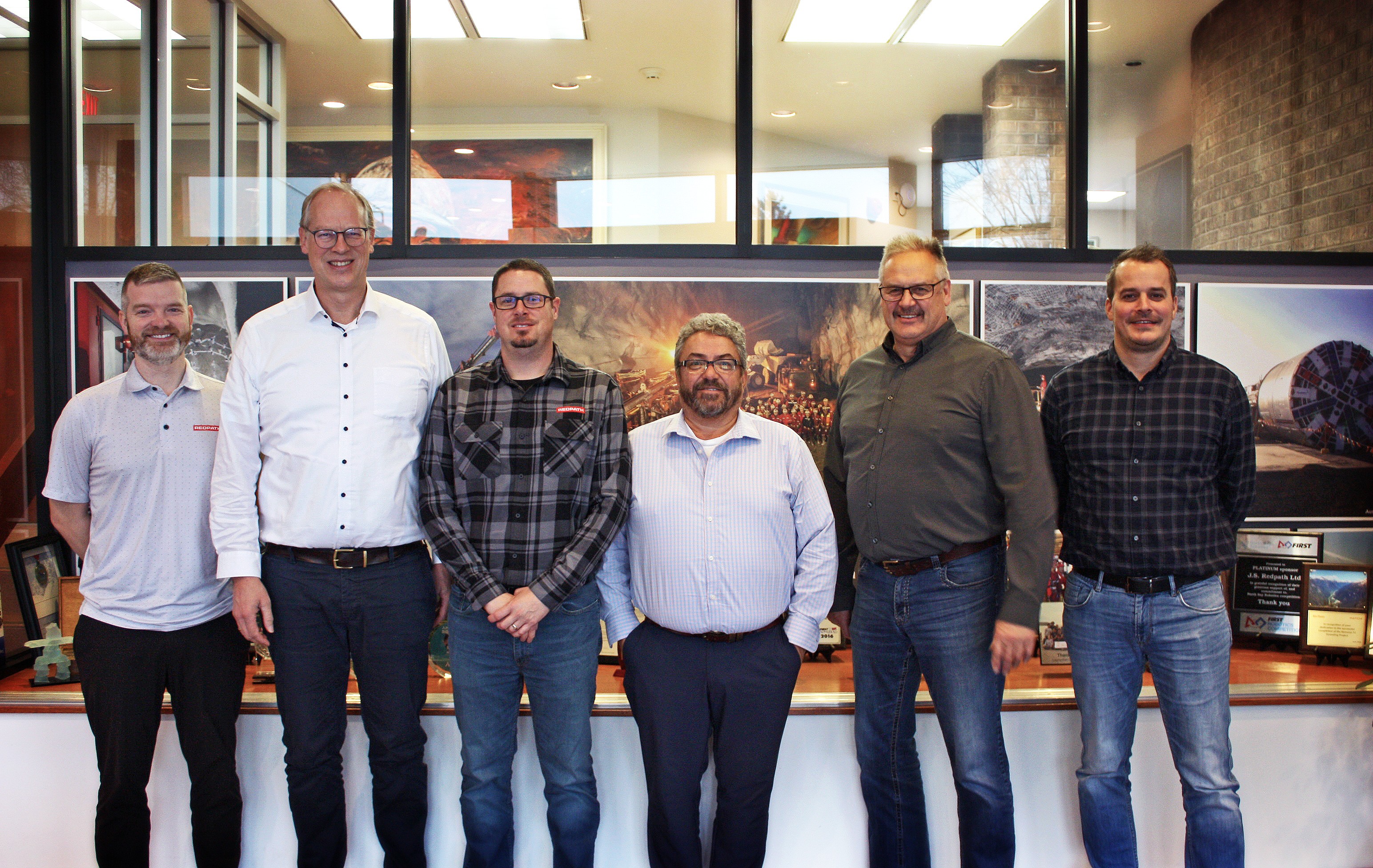 Six smiling representatives from Redpath and Herrenknecht pose with mining photos in the background.